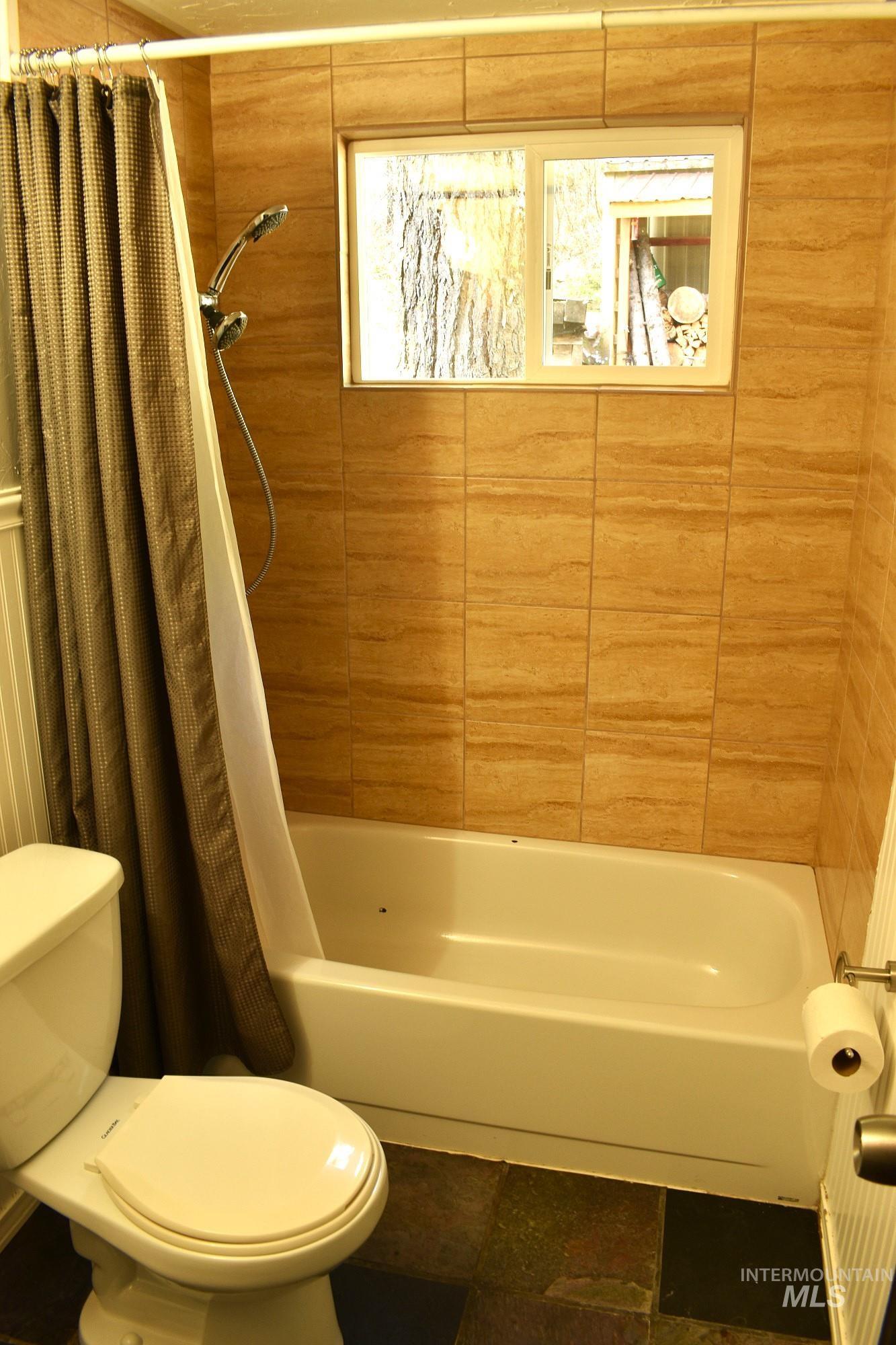 Bathroom featuring a white porcelain tub, a mounted showerhead, a toilet, a window, and earth-toned tile surround