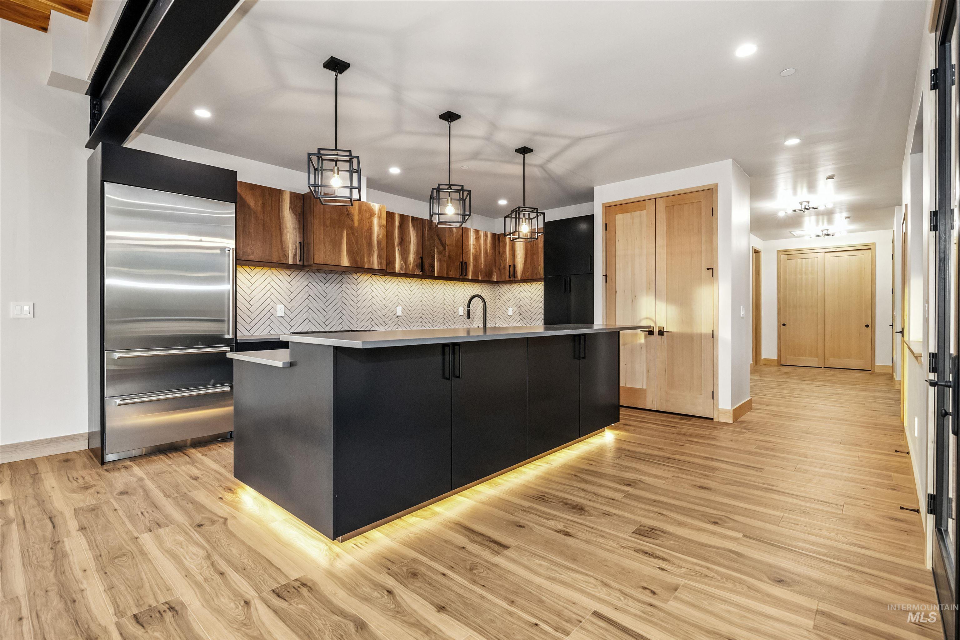 Kitchen with backsplash, a center island with sink, hanging light fixtures, built in fridge, and light wood-type flooring