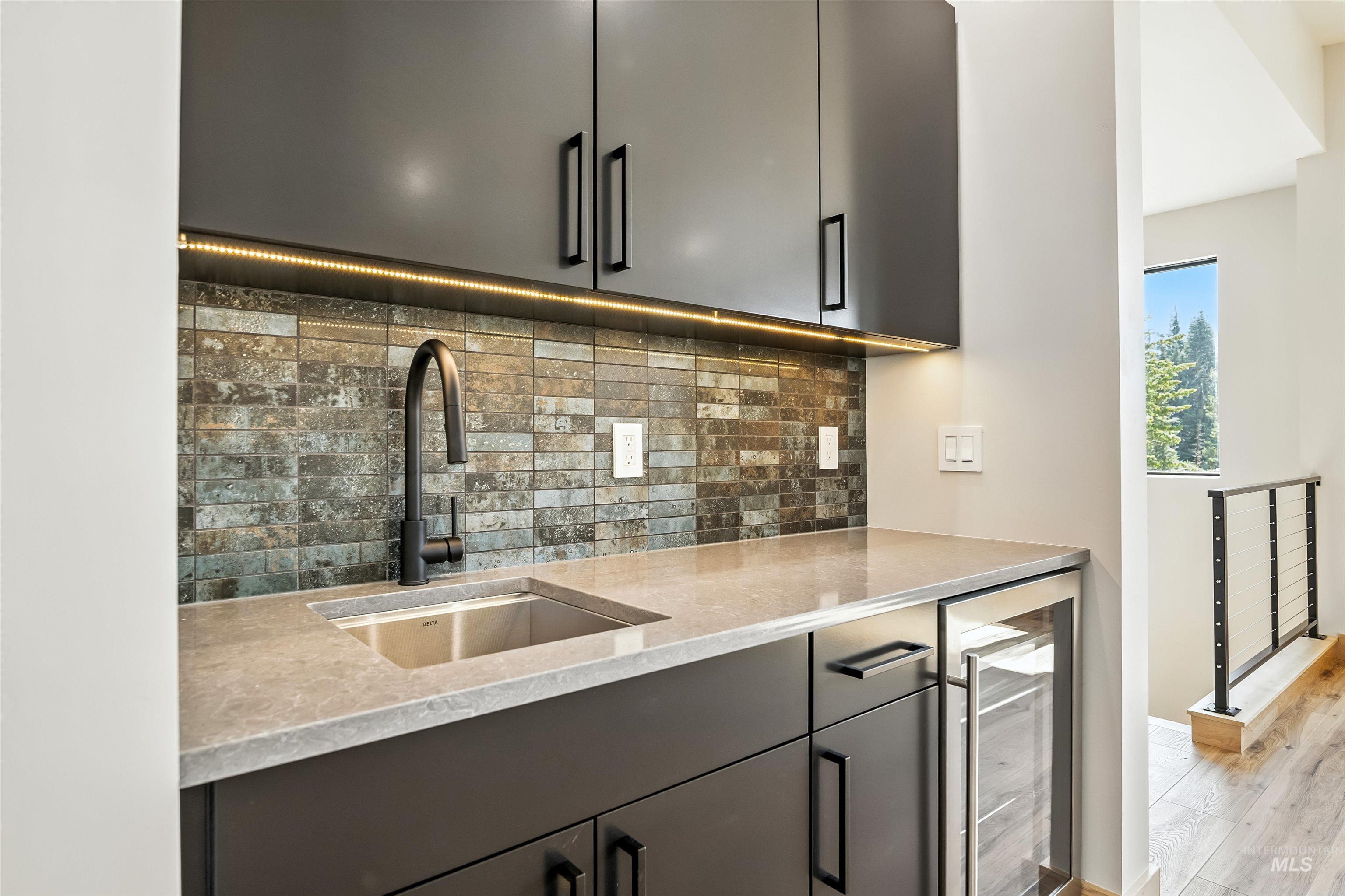 Kitchen with beverage cooler, backsplash, light stone countertops, gray cabinetry, and light wood finished floors