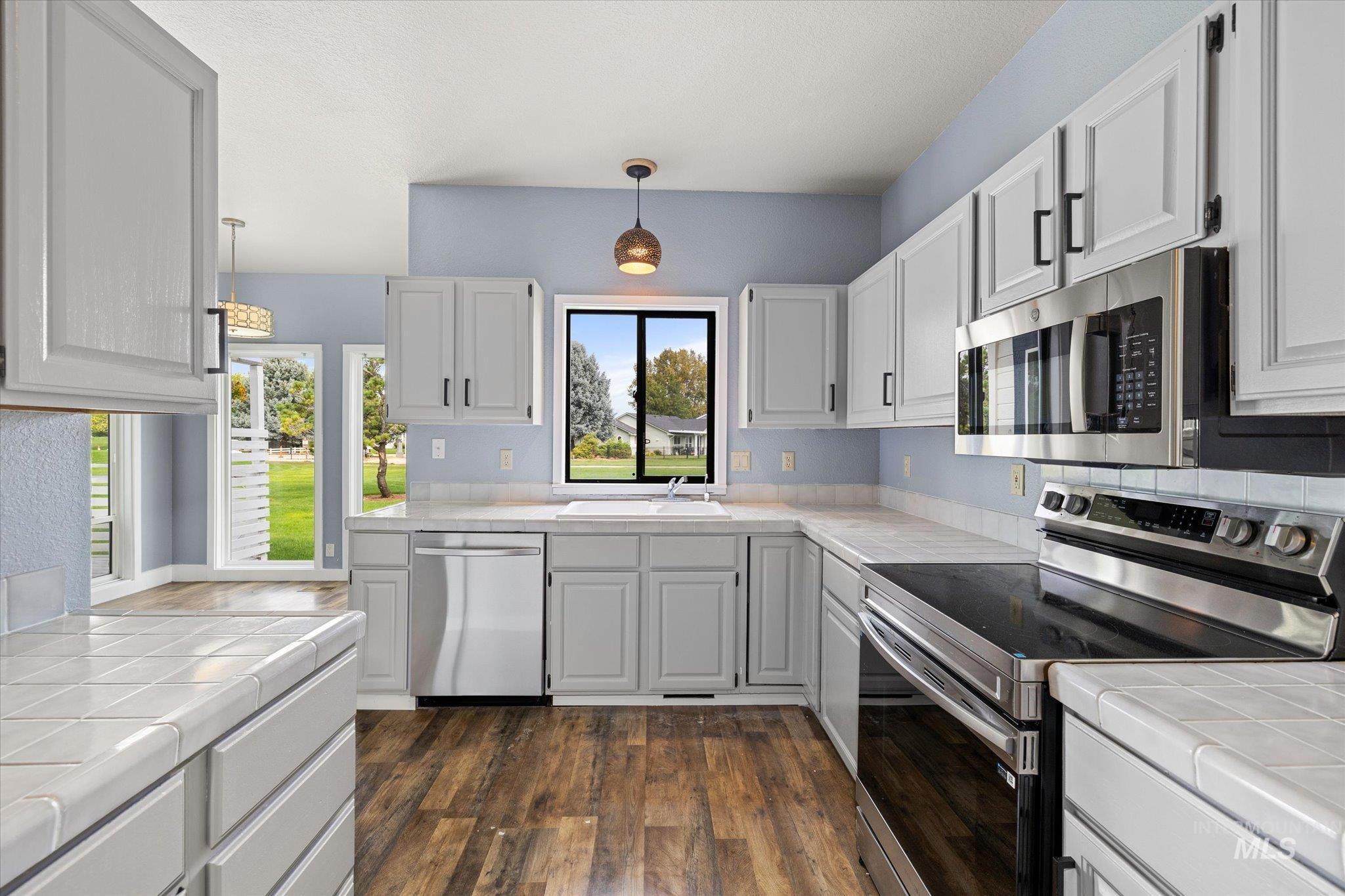 Kitchen with tile counters, appliances with stainless steel finishes, dark wood finished floors, decorative light fixtures, and white cabinetry