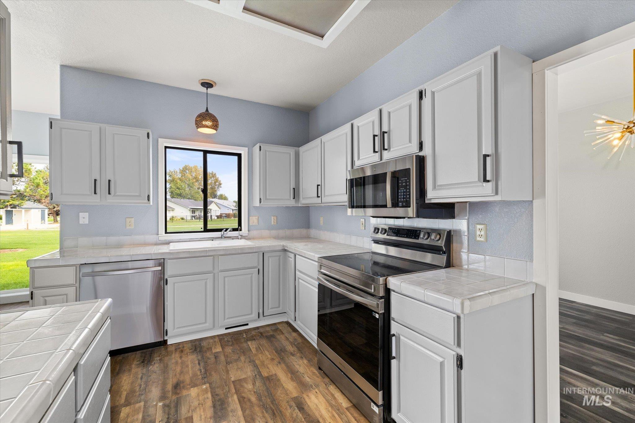 Kitchen featuring stainless steel appliances, dark wood-style floors, hanging light fixtures, and tile countertops