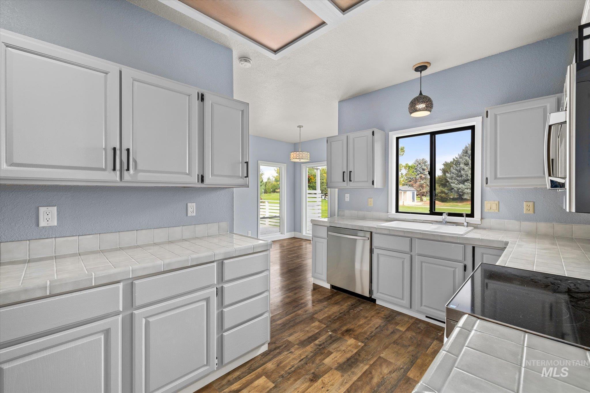 Kitchen with tile counters, a textured wall, dark wood-style floors, appliances with stainless steel finishes, and decorative light fixtures