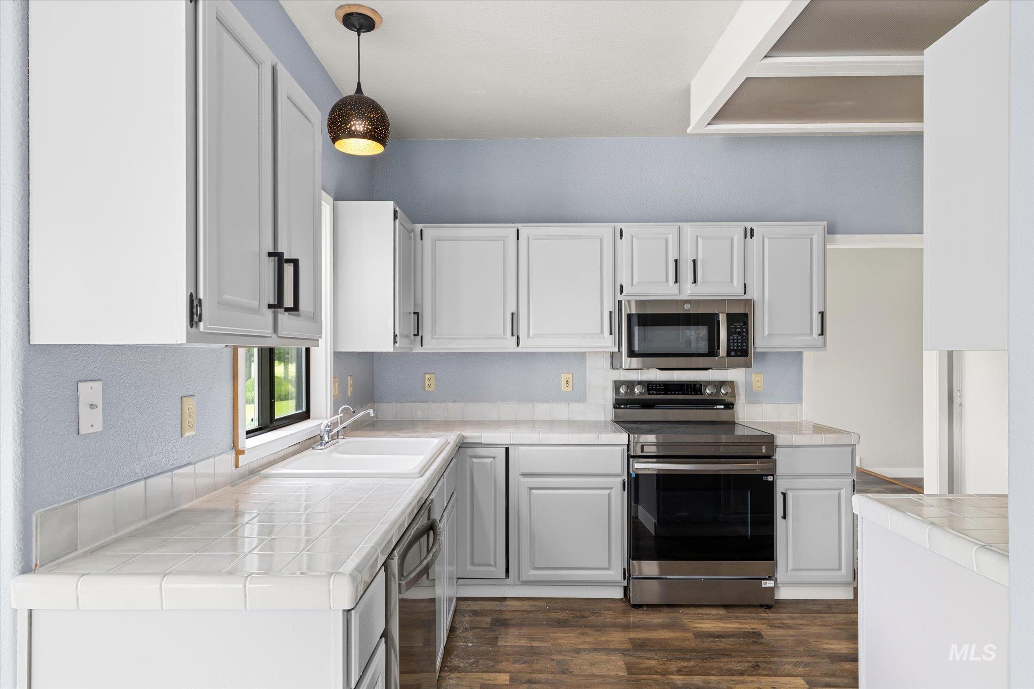 Kitchen with tile counters, stainless steel appliances, dark wood-style floors, and white cabinetry