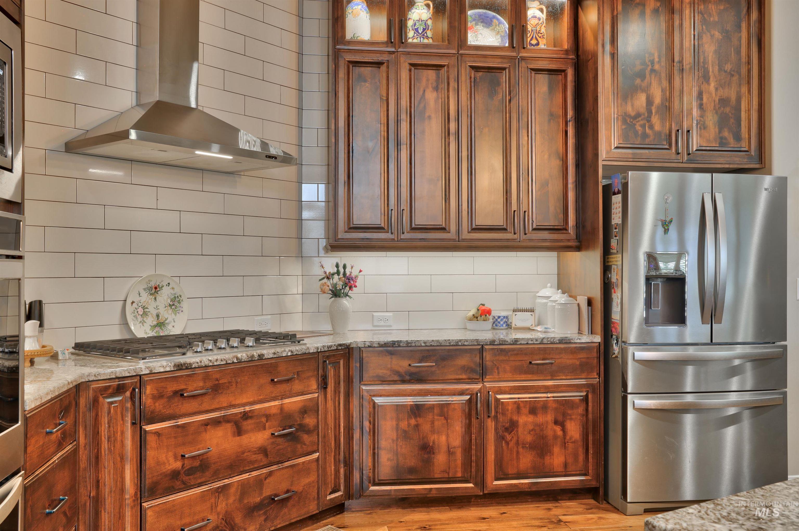 Kitchen with stainless steel appliances, wall chimney exhaust hood, light stone counters, glass insert cabinets, and tasteful backsplash