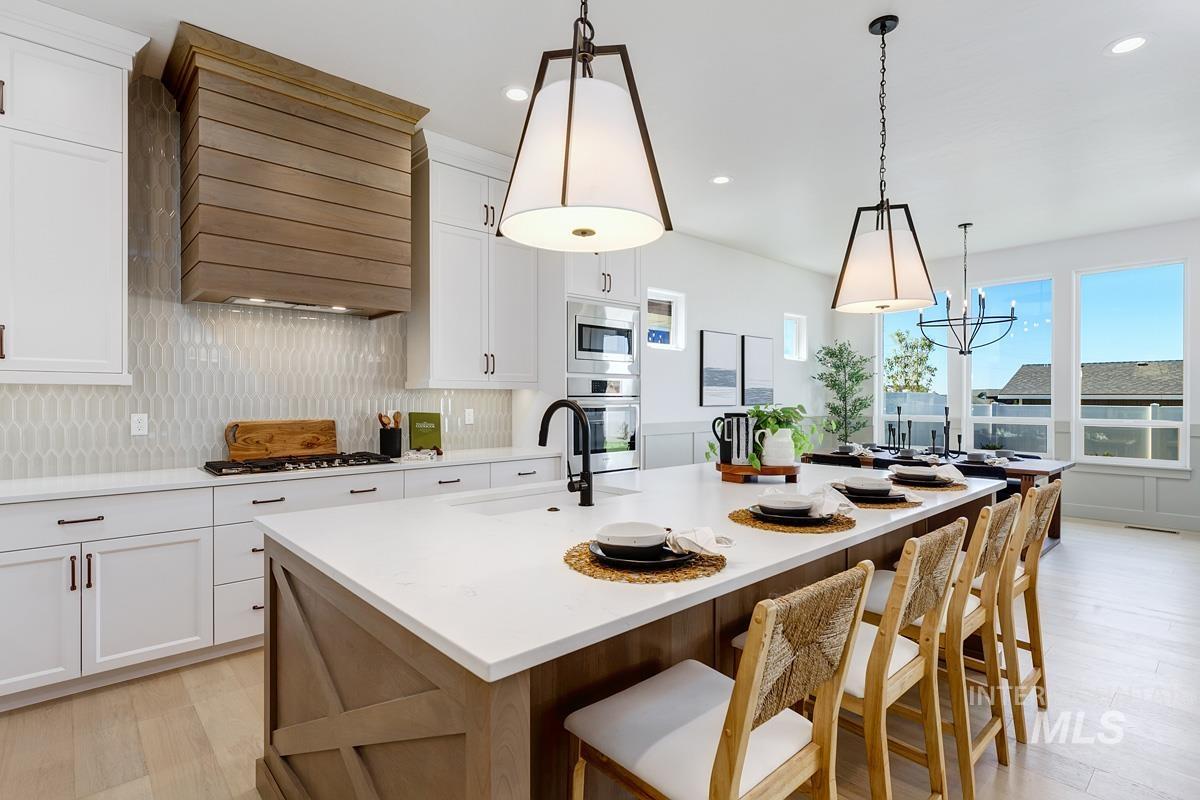 Kitchen with light wood finished floors, white cabinetry, an island with sink, hanging light fixtures, and light stone countertops