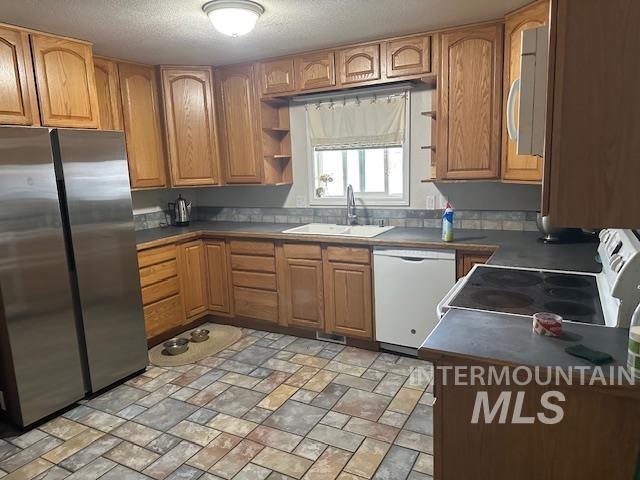 Kitchen with freestanding refrigerator, dark countertops, brown cabinets, white dishwasher, and a textured ceiling