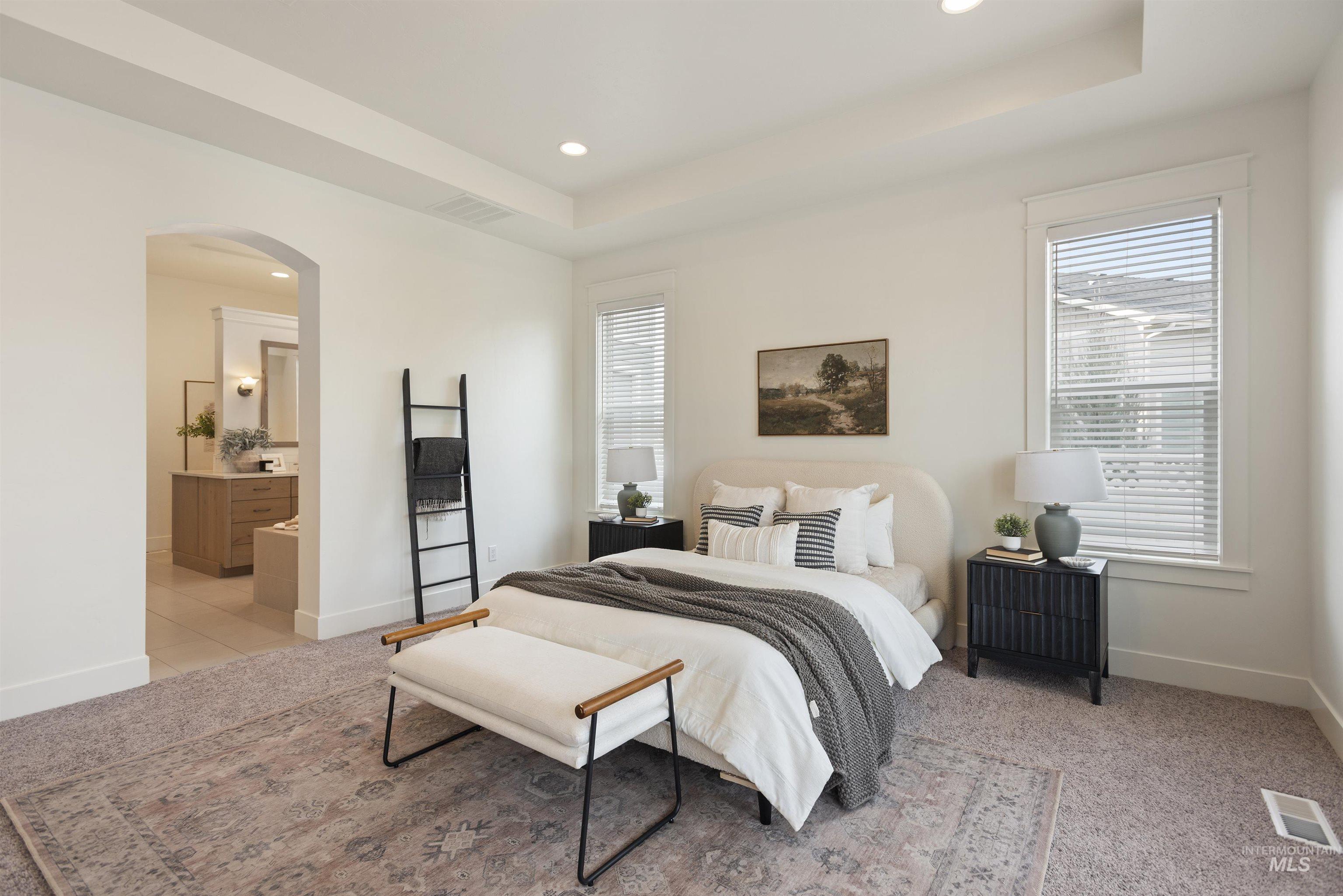 Bedroom with arched walkways, a tray ceiling, recessed lighting, light colored carpet, and ensuite bath