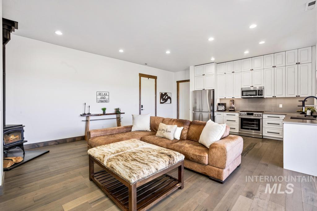 Open-concept living space featuring wood-finish flooring, a freestanding wood-burning stove, recessed lighting, and a kitchen with white shaker cabinetry, stainless steel appliances, and a peninsula with a dark countertop