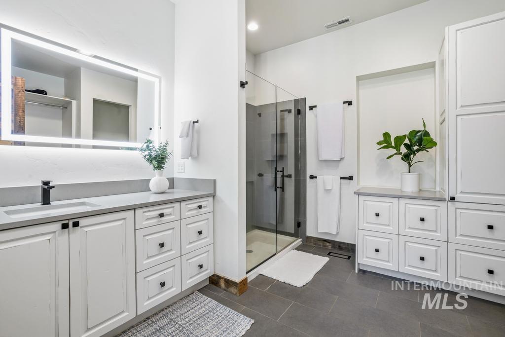 Spacious bathroom featuring a frameless glass shower enclosure, a vanity with an integrated sink and light-up mirror, extensive white cabinetry with black hardware, stone countertops, and large format floor tiling