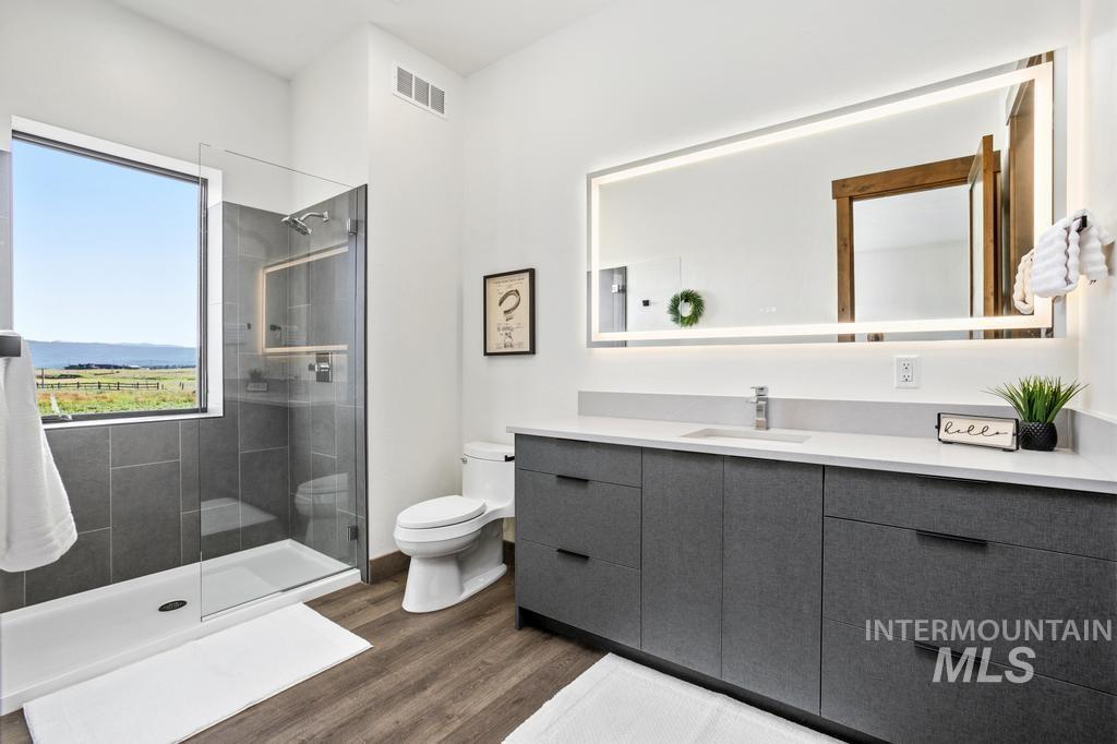 Modern bathroom featuring a spacious glass-enclosed shower, a large window, contemporary vanity with a white countertop, wood-finish flooring, and a backlit mirror