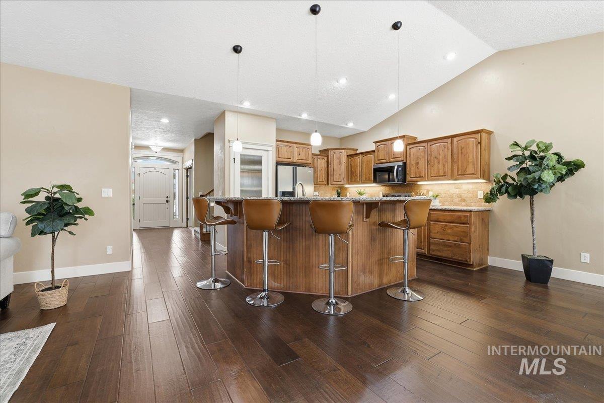Kitchen with brown cabinetry, hanging light fixtures, light stone counters, an island with sink, and a breakfast bar