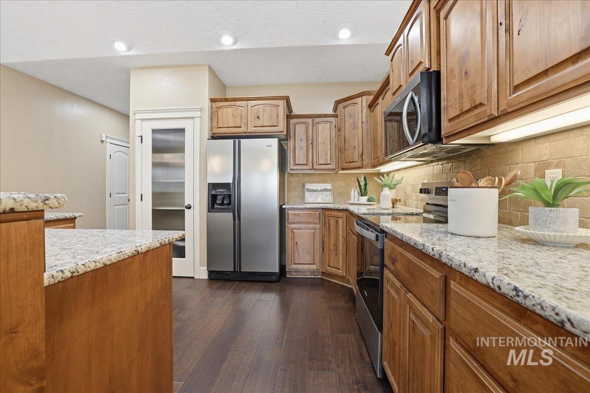 Kitchen featuring light stone counters, appliances with stainless steel finishes, dark wood-style flooring, brown cabinets, and backsplash