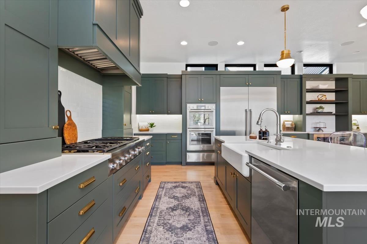 Kitchen with light wood-style floors, backsplash, hanging light fixtures, stainless steel appliances, and an island with sink