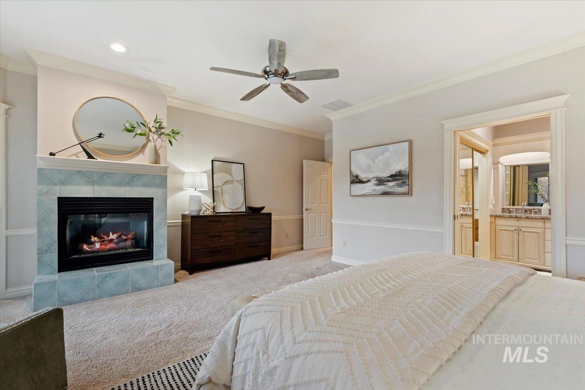 Carpeted bedroom featuring a tiled fireplace, ornamental molding, a ceiling fan, and ensuite bath