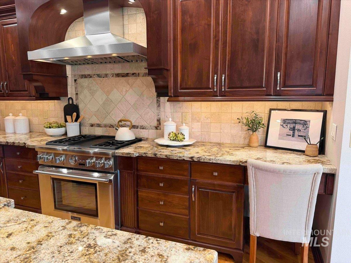 Kitchen with light stone countertops, exhaust hood, stainless steel stove, backsplash, and dark wood finish cabinetry