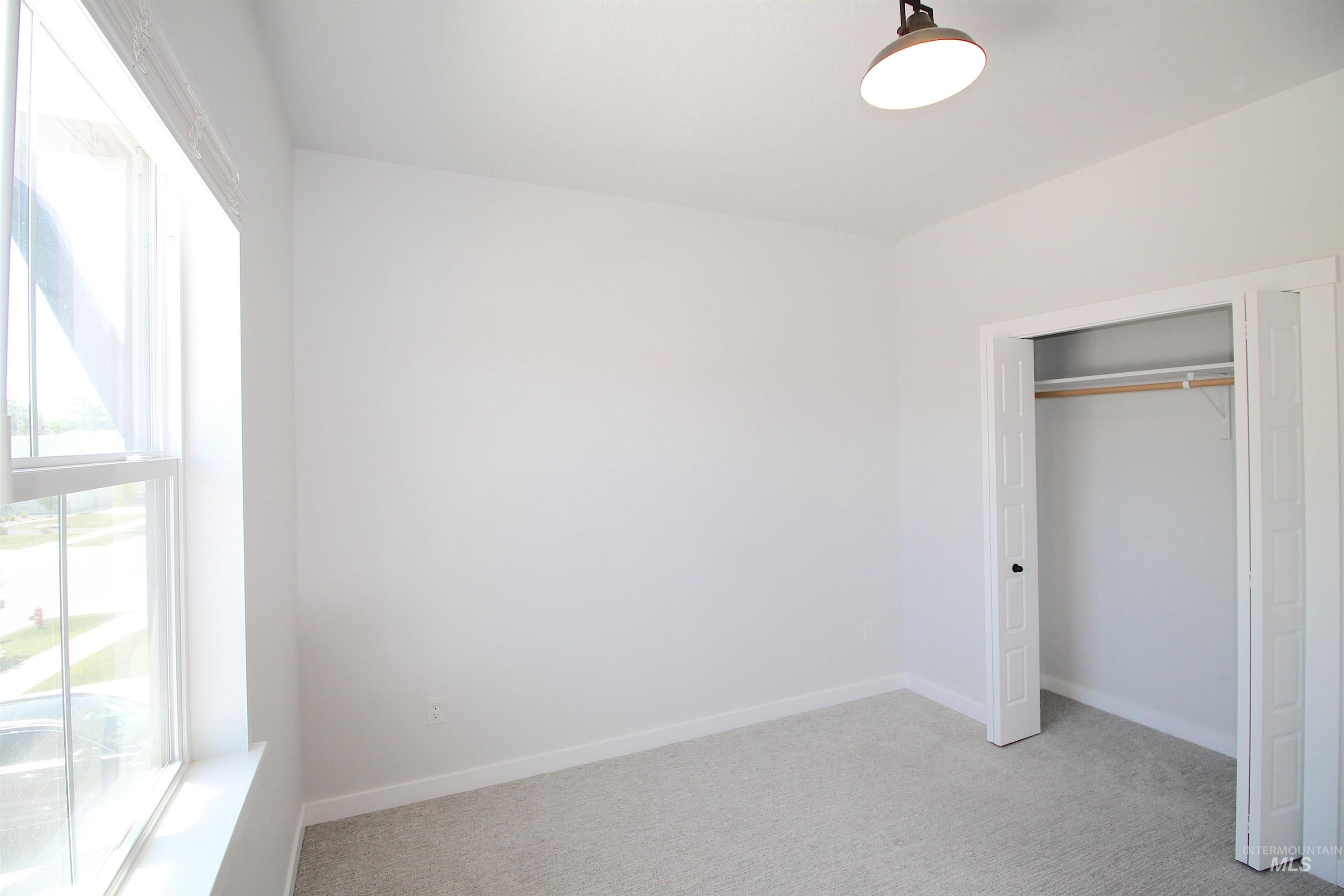 Bright room with a large window, neutral carpet flooring, white walls, and a bi-fold closet featuring a shelf and hanging rod