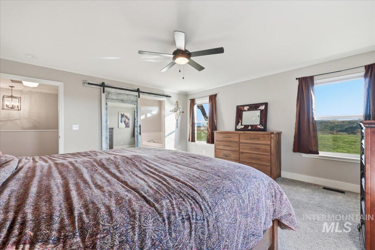 Bedroom with a barn door, crown molding, ensuite bath, light carpet, and a ceiling fan