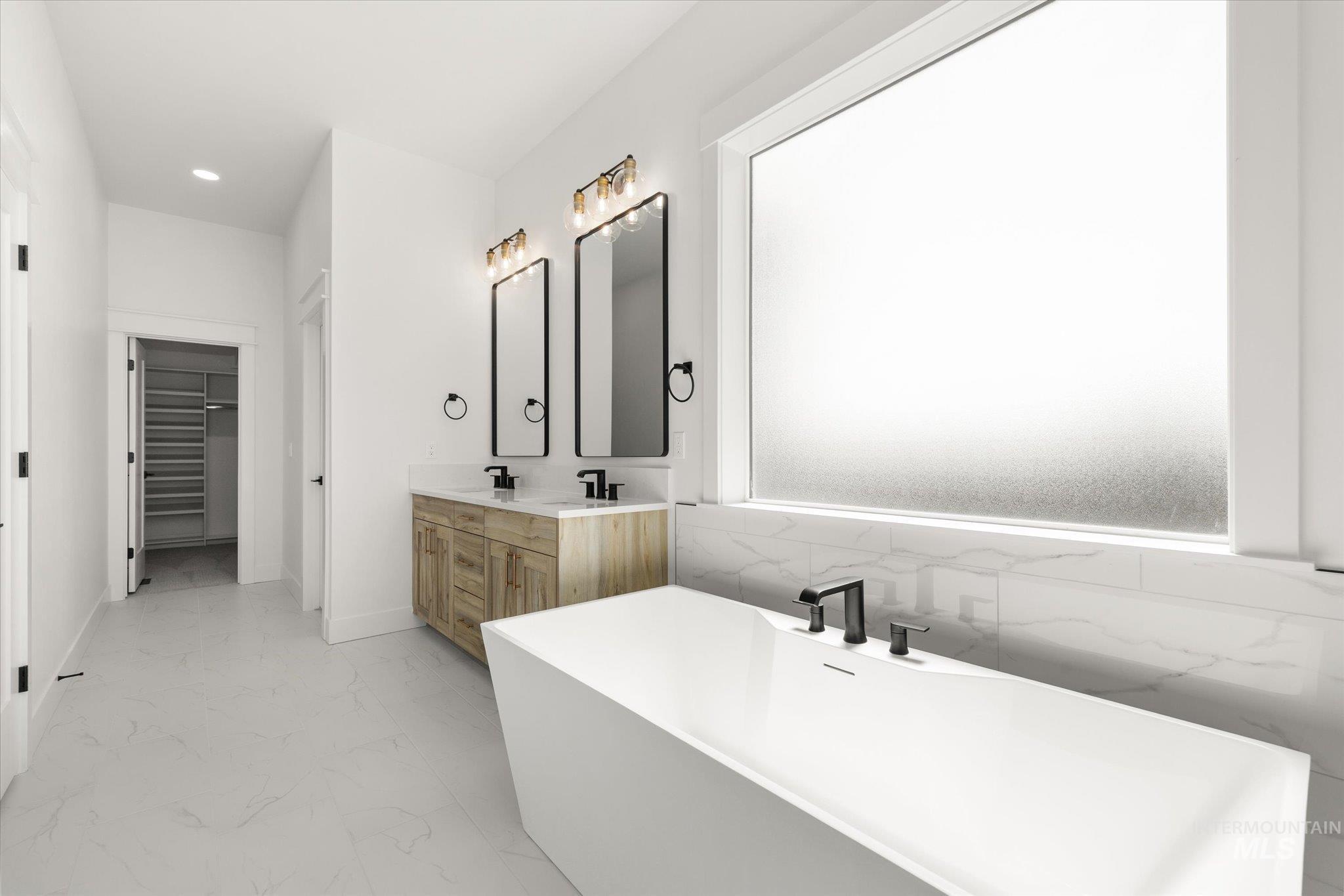 Bathroom featuring double vanity, a walk in closet, a freestanding tub, light marble finish flooring, and recessed lighting