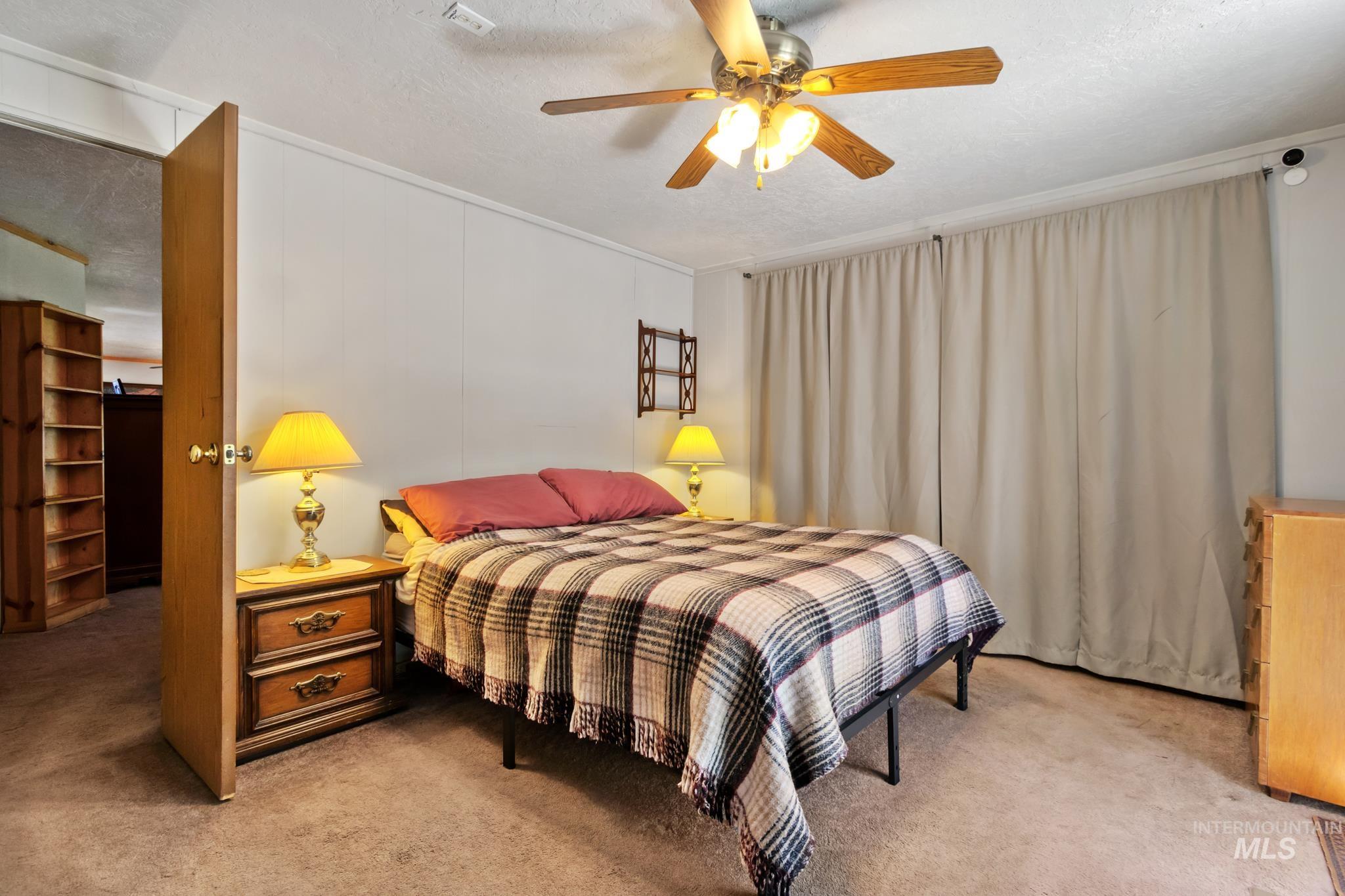 Carpeted room featuring a ceiling fan with integrated lighting, wood-finish wall paneling, and a large window opening with light-colored drapes