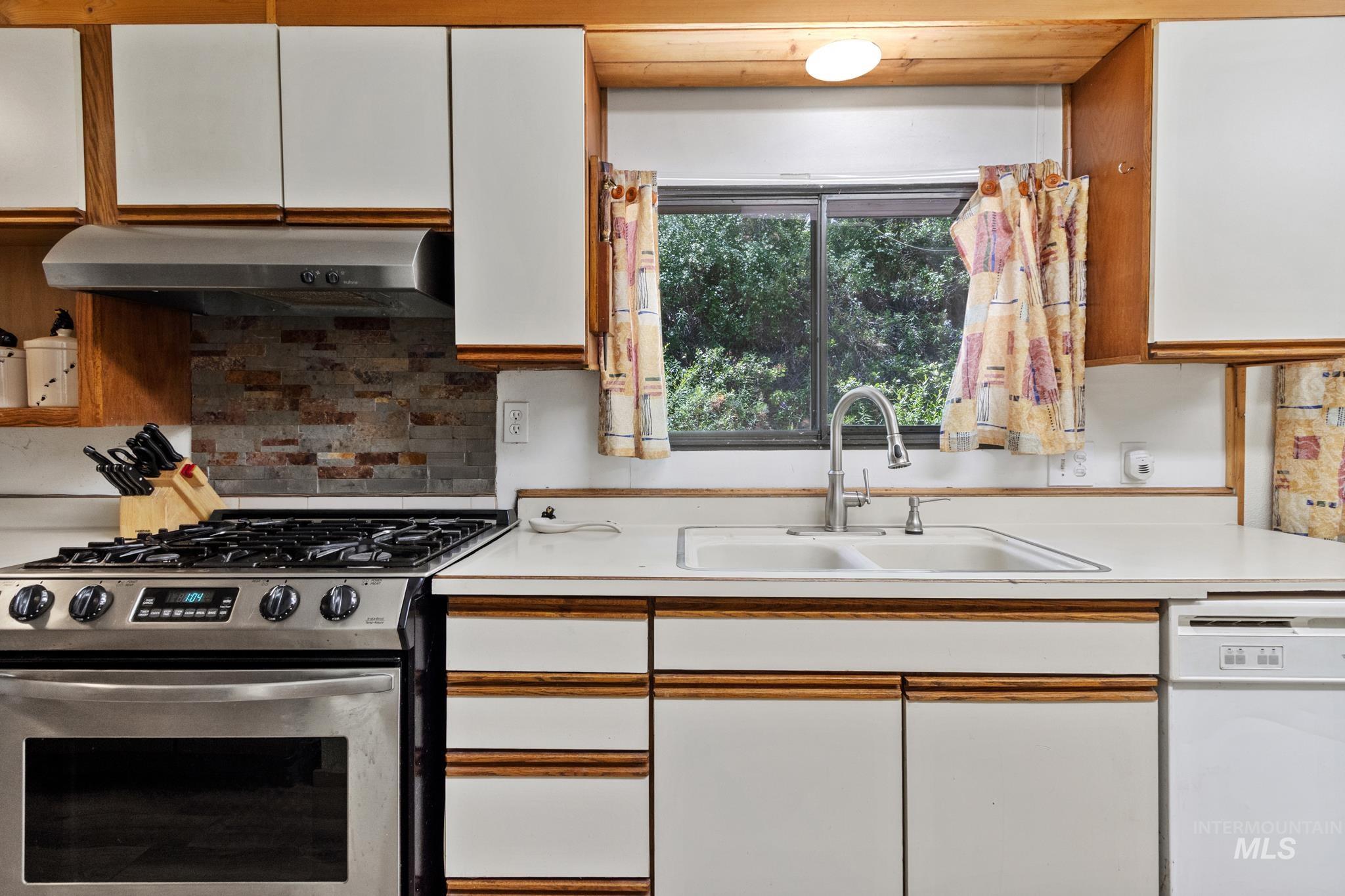 Kitchen featuring white cabinetry with wood trim, a stainless steel range with a gas cooktop, a coordinating stainless steel range hood, stone tile backsplash, and a double basin sink
