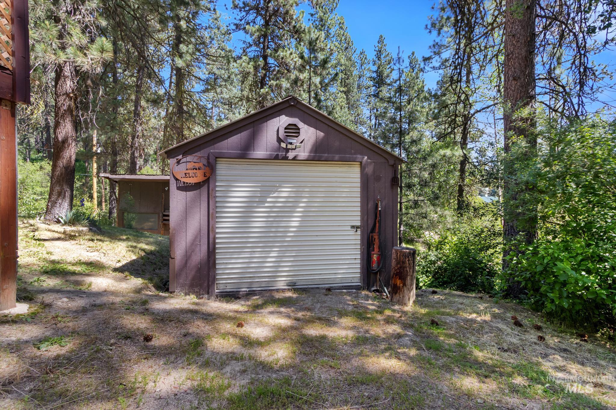 Detached brown storage shed featuring a rolling garage door and side awning structure, surrounded by mature trees