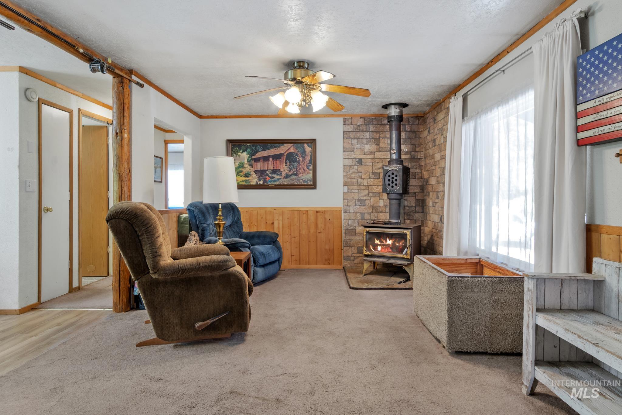 Living area featuring a floor-to-ceiling stone accent wall with a wood-burning stove, light wood wainscoting, and a ceiling fan with integrated lighting