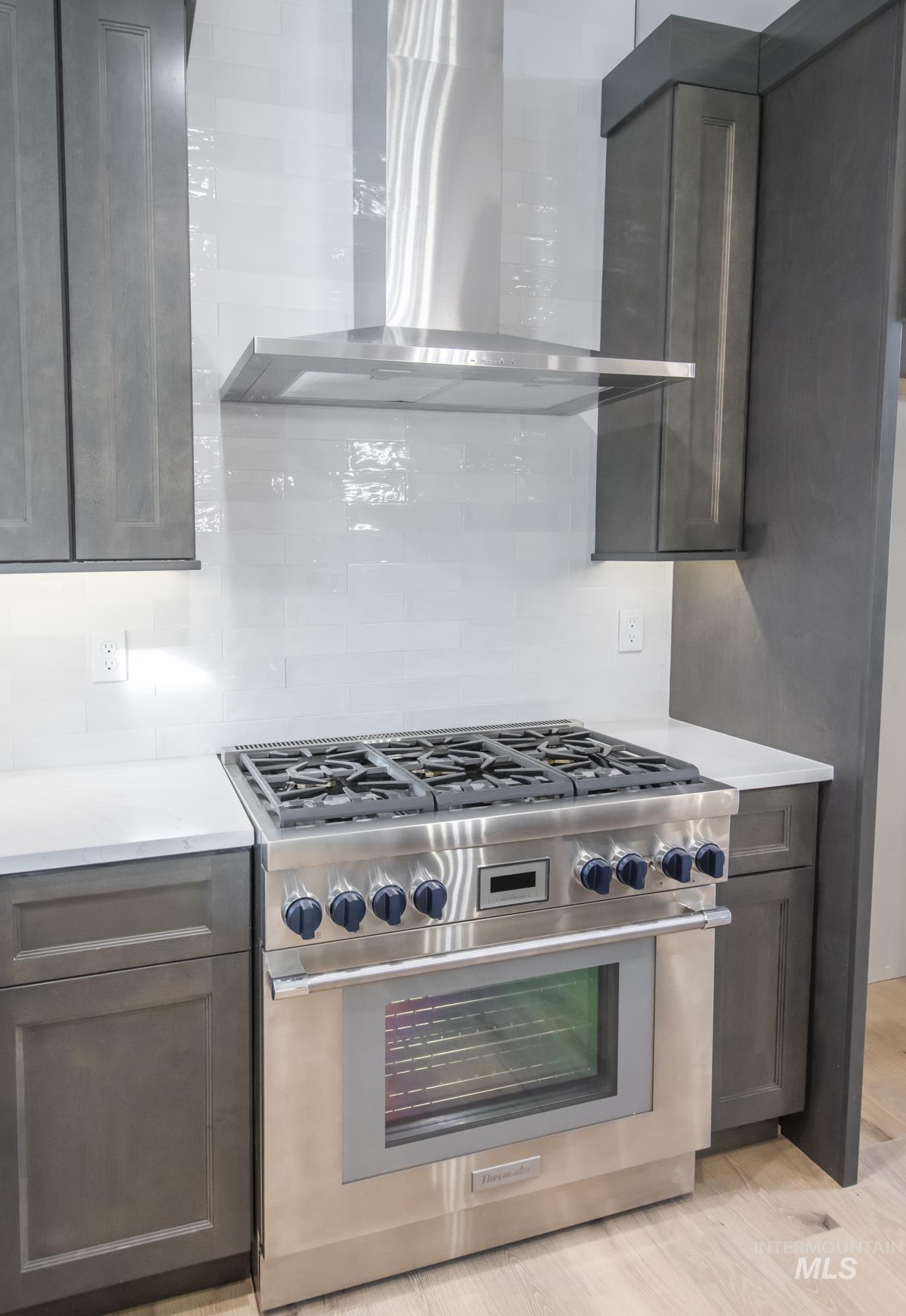 Kitchen with wall chimney exhaust hood, stainless steel Thermador gas range, quartz countertops, light wood finished floors, and tile backsplash