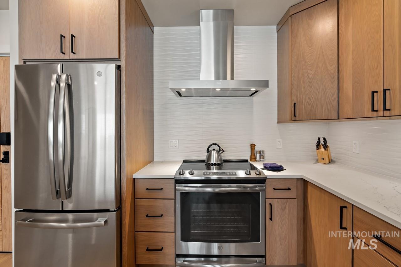 Kitchen with appliances with stainless steel finishes, wall chimney exhaust hood, backsplash, light countertops, and brown cabinets