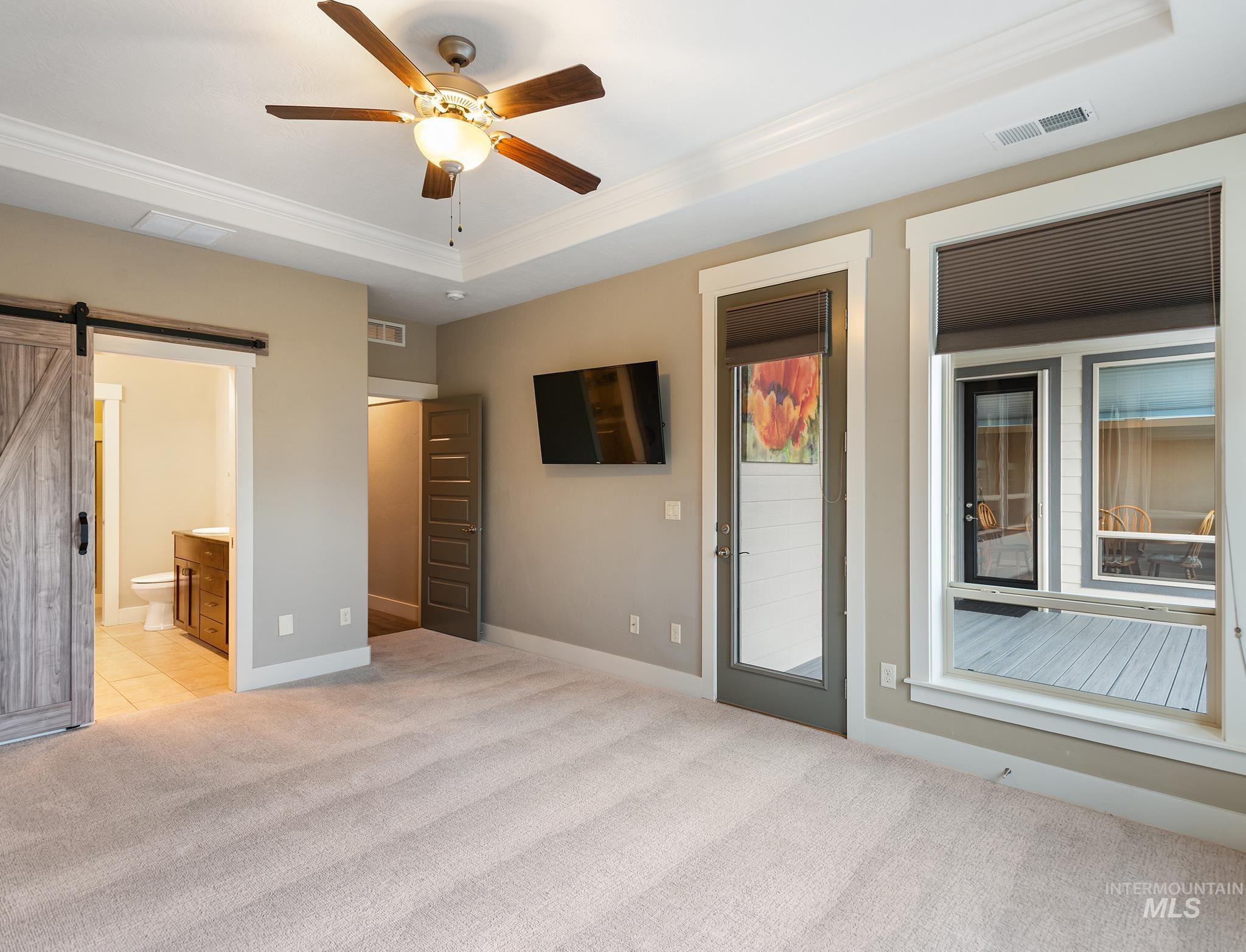 Unfurnished bedroom featuring a barn door, a tray ceiling, crown molding, ensuite bath, and light colored carpet