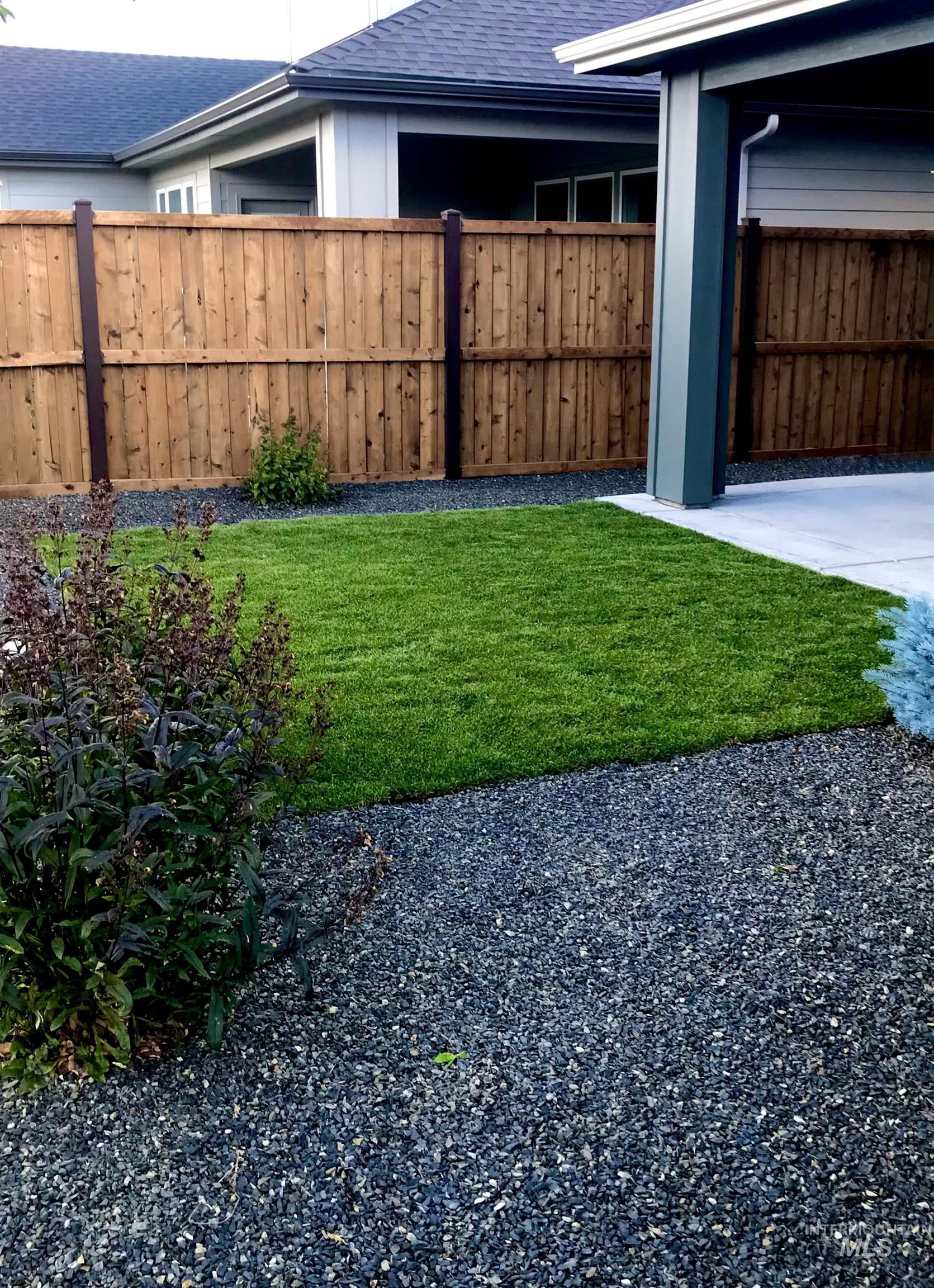 Landscaped exterior featuring a private wood fence, a section of manicured lawn, a concrete patio area, dark gravel ground cover, and planted shrubbery
