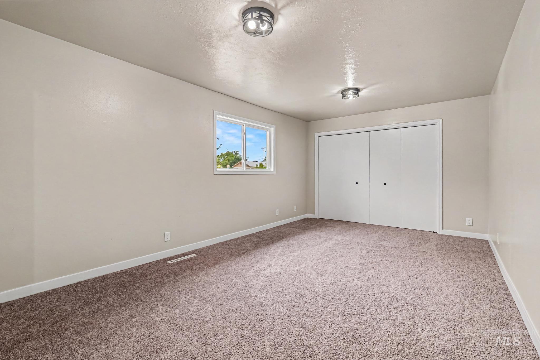 Spacious room featuring neutral-toned walls, plush carpet flooring, a large window, two flush-mount ceiling lights, and bi-fold closet doors