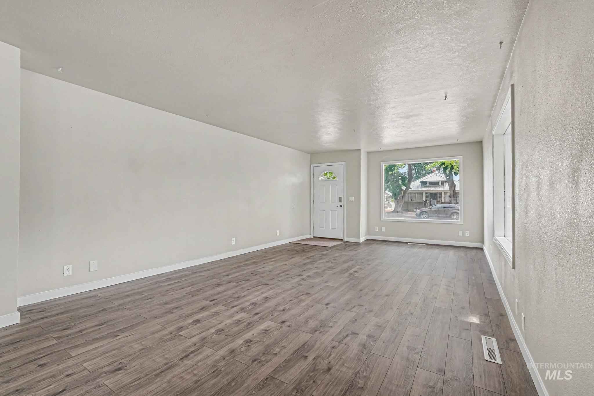Spacious living area featuring wood-finish flooring, light grey wall paint, white baseboards, and a prominent front-facing window