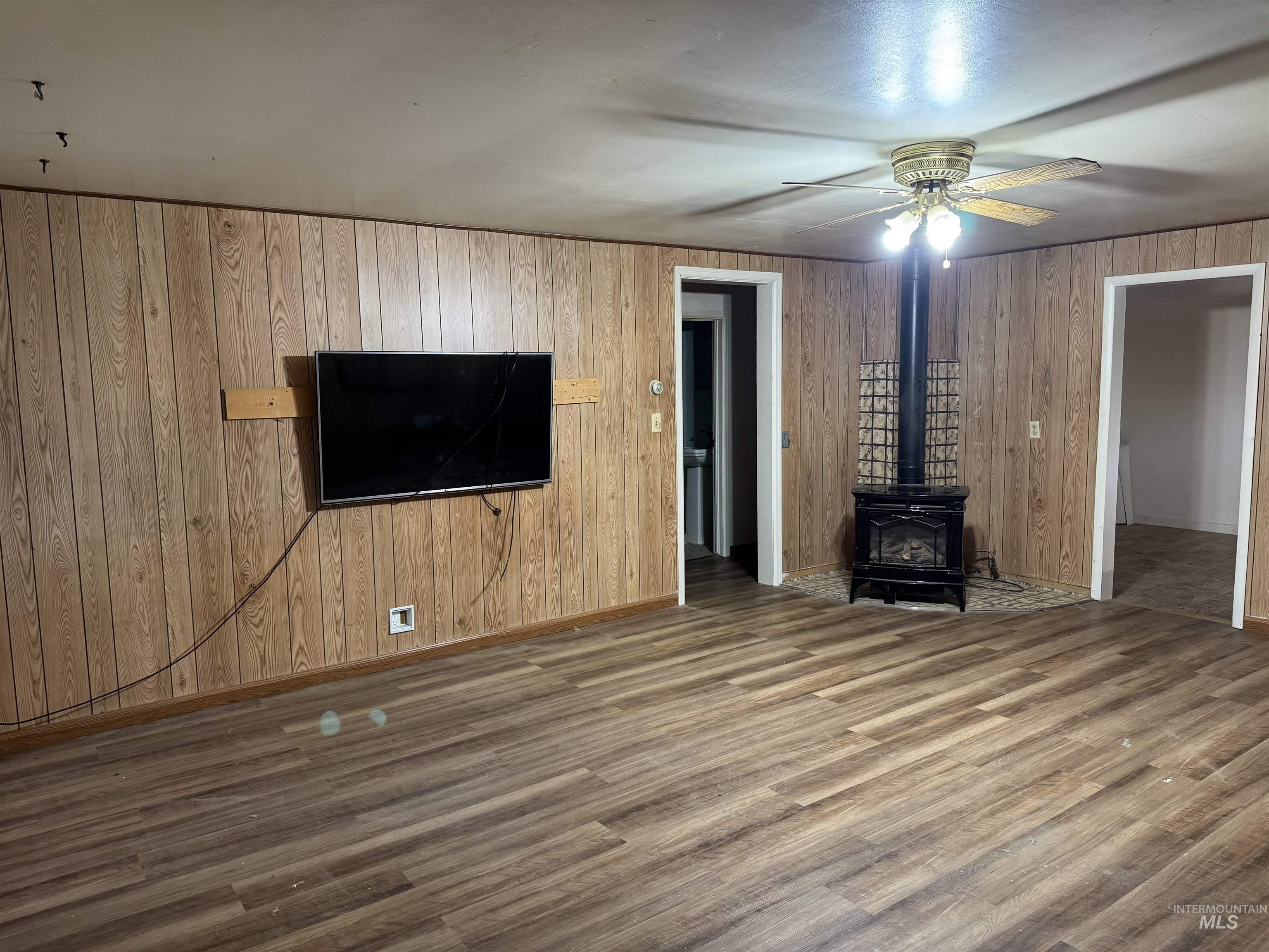 Unfurnished living room featuring a wood stove, wooden walls, wood finished floors, and ceiling fan