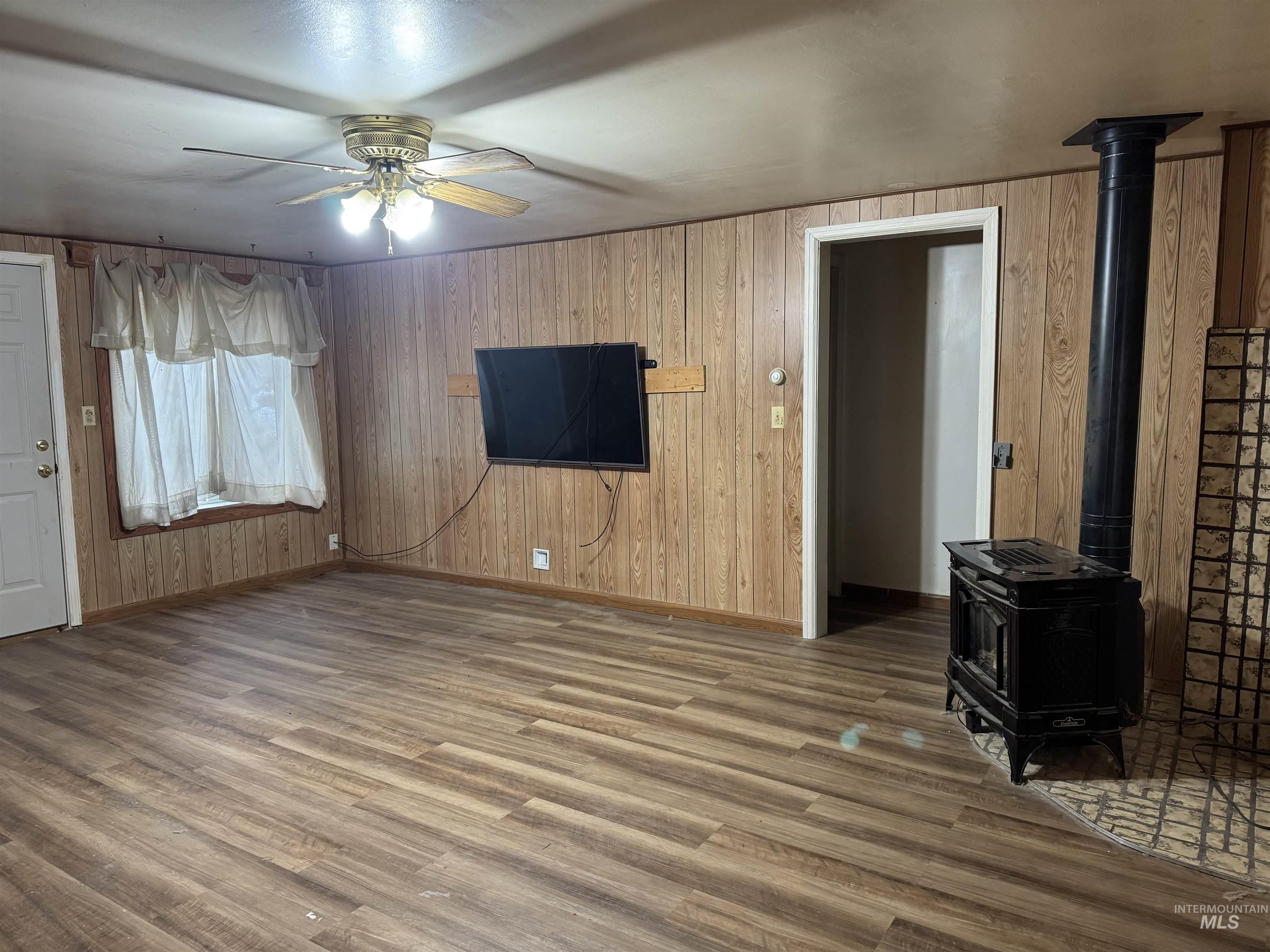Unfurnished living room with a wood stove, wood finished floors, a ceiling fan, and wooden walls