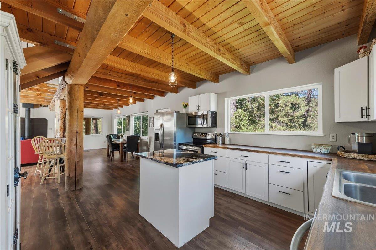Open-concept kitchen featuring wood-finish flooring, white cabinetry with black hardware, a center island with a stone countertop, a stainless steel appliance suite, and a wood plank vaulted ceiling with exposed beams