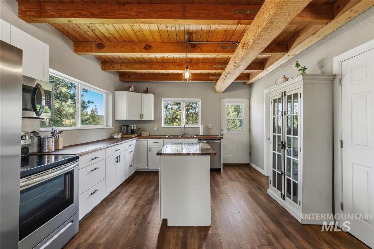 Kitchen featuring exposed wood beam ceilings, wood-finish flooring, white cabinetry with dark hardware, stainless steel appliances, and a central island with a stone countertop