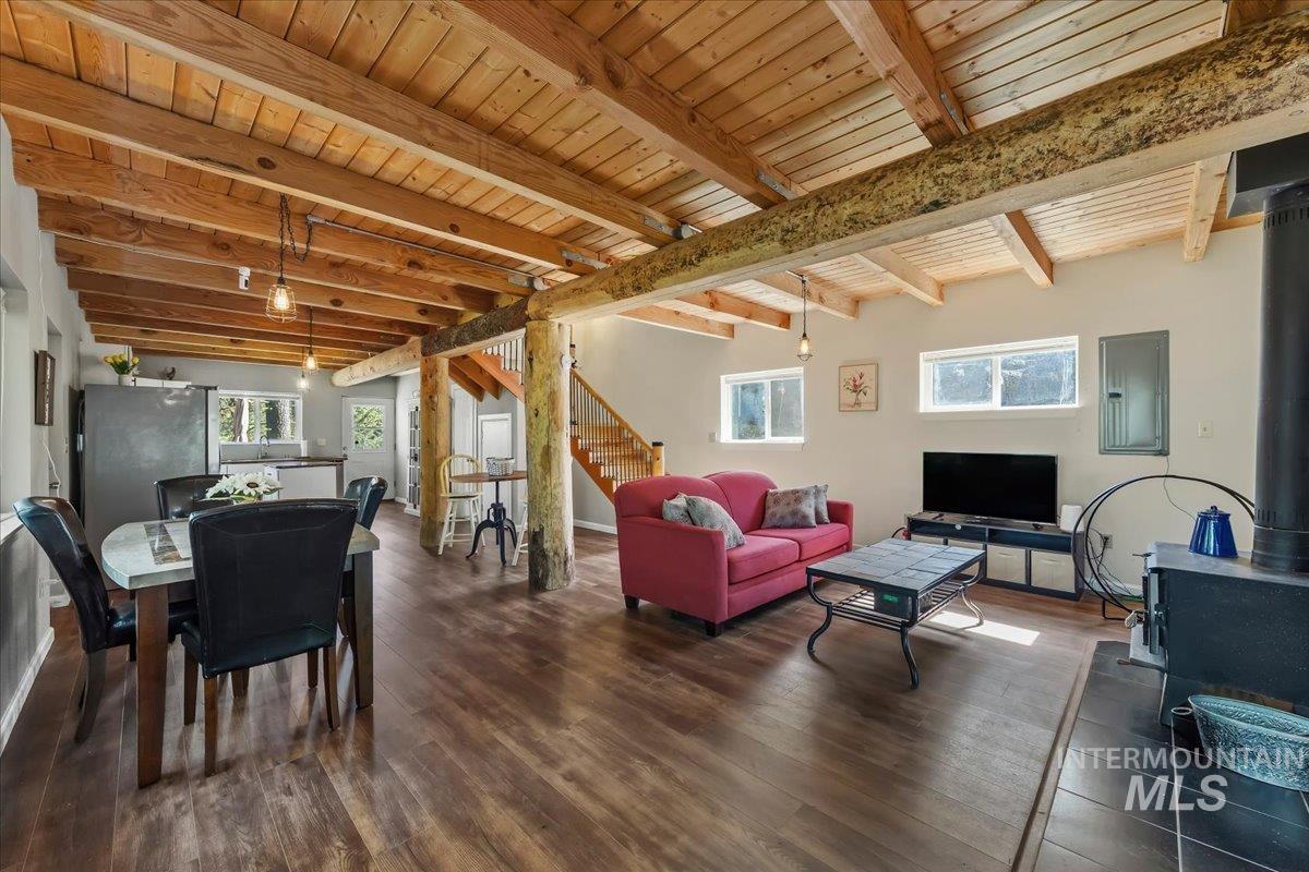 Open-concept living area featuring exposed wood-beam ceilings, wood-finish flooring, a built-in wood-burning stove with tiled hearth, a visible staircase with wood railing, and multiple windows for abundant natural light