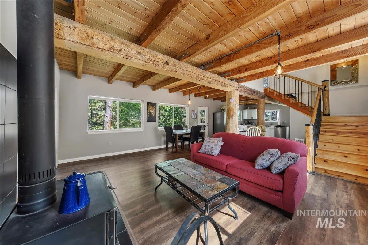 Spacious living area featuring exposed wood beam ceilings, wood-finish flooring, multiple windows, a wood stove, and a visible staircase with wrought iron balusters