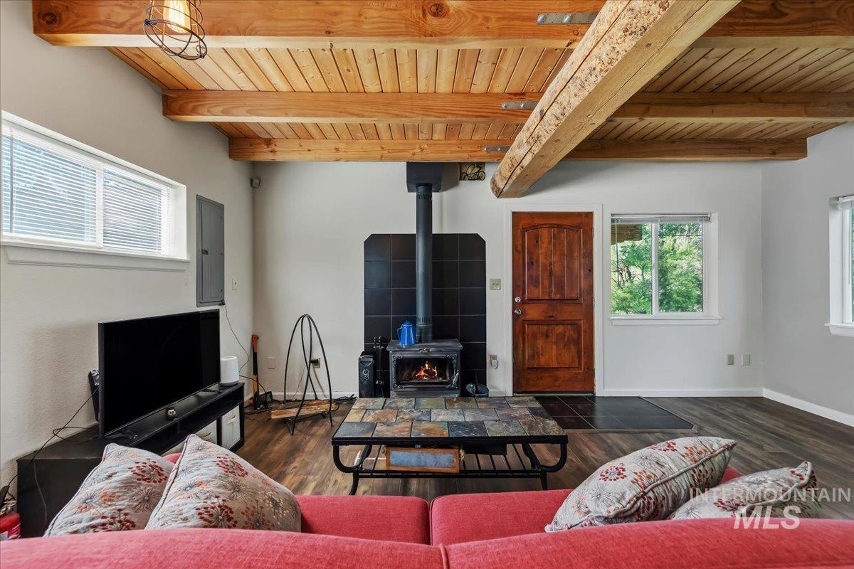 Living area featuring wood-finish flooring, exposed wood beam ceiling, a black tile wall with a built-in wood stove, and a solid wood entry door