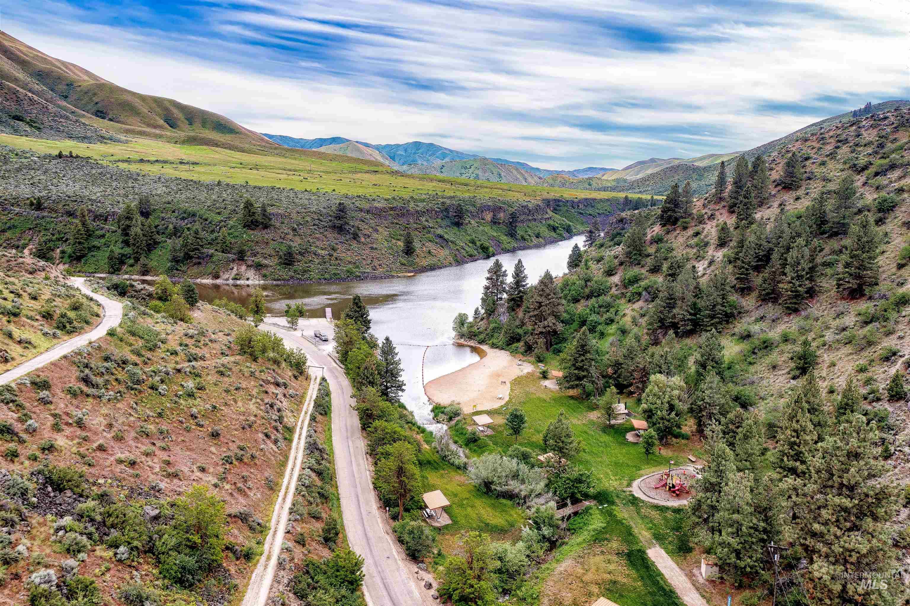 Winding paved roads leading to a riverfront area featuring a boat launch, sandy beach, and a grassy recreational space with mature trees