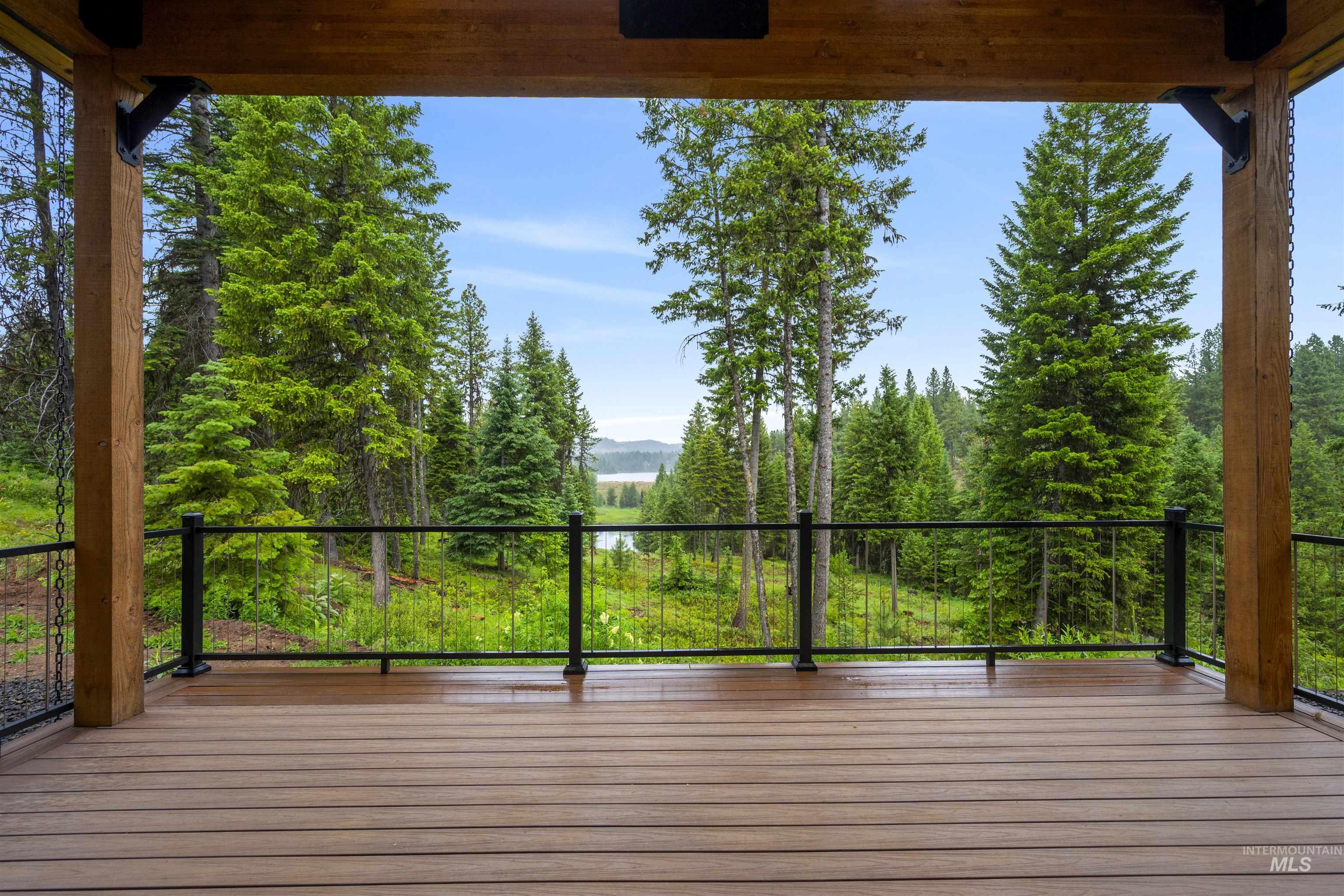 Covered porch featuring wood-finish decking, black metal railing, exposed timber beams, and a natural wooded backdrop with a distant body of water