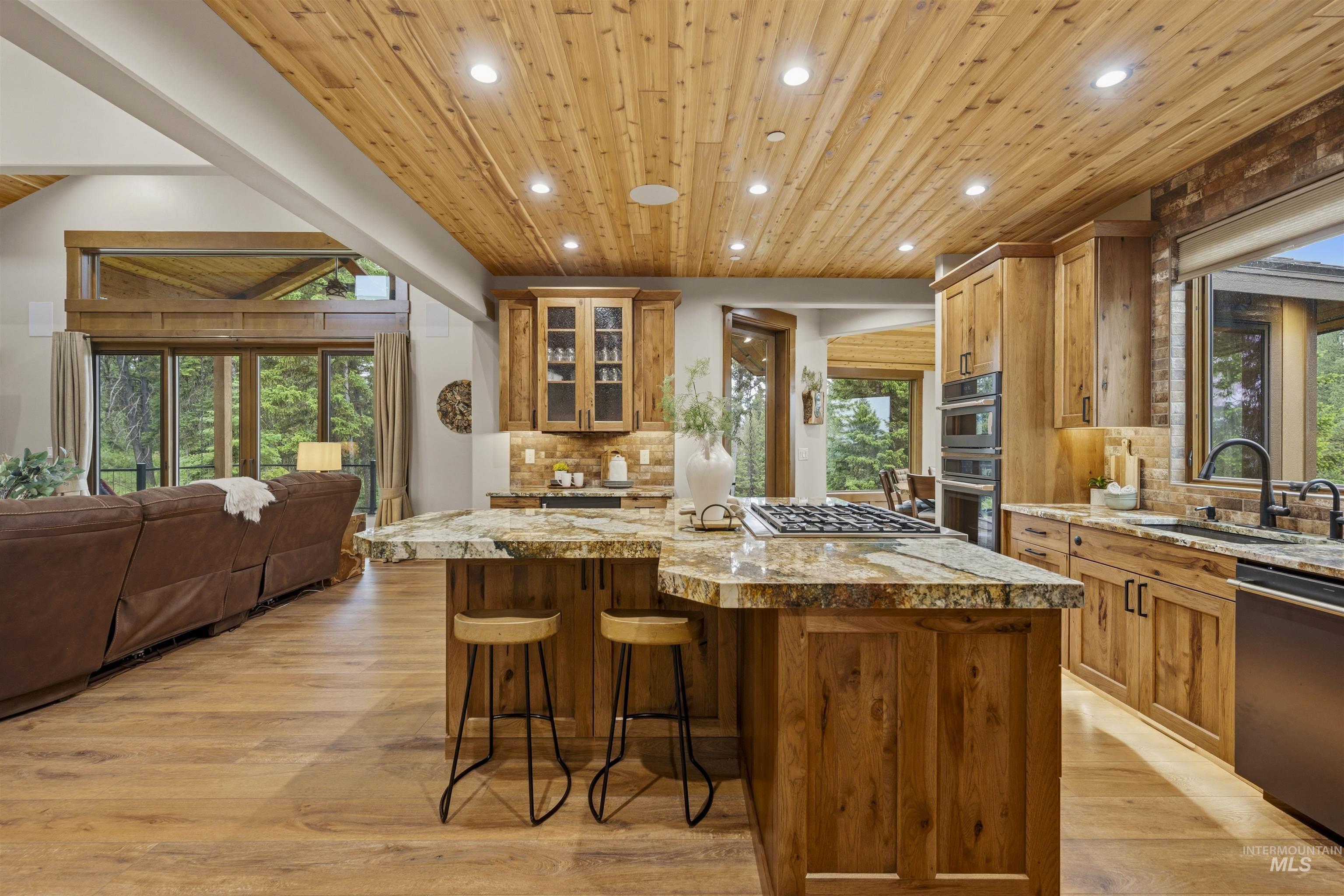 Open-concept kitchen featuring wood-finish cabinetry, a large island with integrated cooktop, stone countertops, and stainless steel built-in appliances