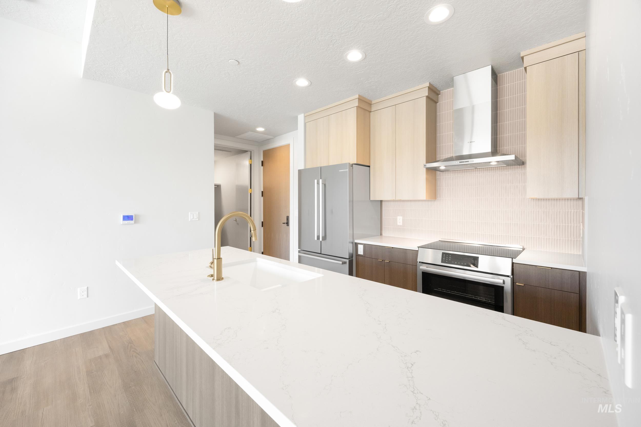 Kitchen featuring modern cabinets, wall chimney exhaust hood, decorative light fixtures, appliances with stainless steel finishes, and light stone countertops