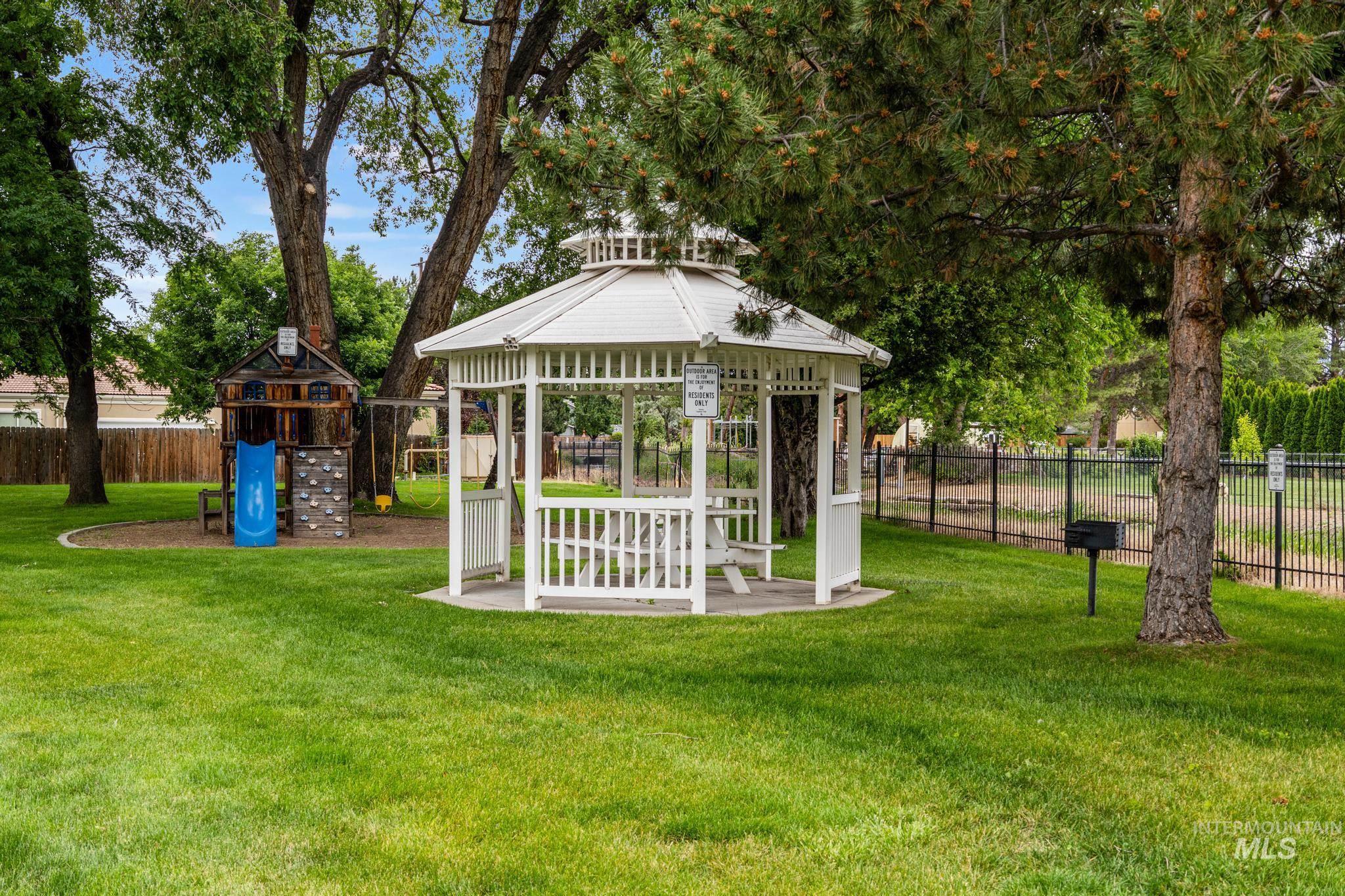Expansive green space featuring a white gazebo with built-in seating, a black metal fence, a play structure with a slide, and mature trees providing natural shade