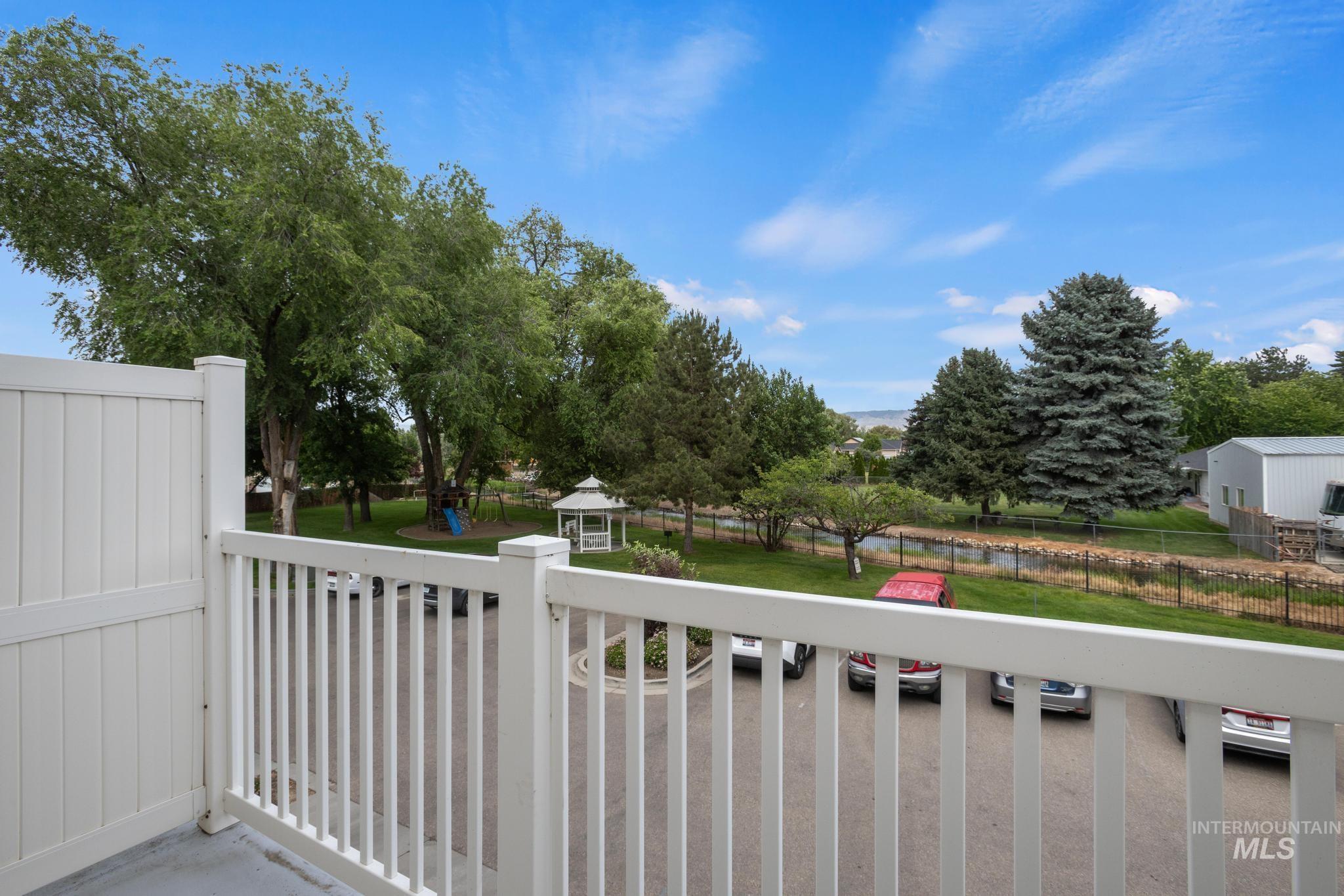 Balcony with white railing and privacy wall overlooks a green space featuring a white gazebo, mature trees, and a distant body of water