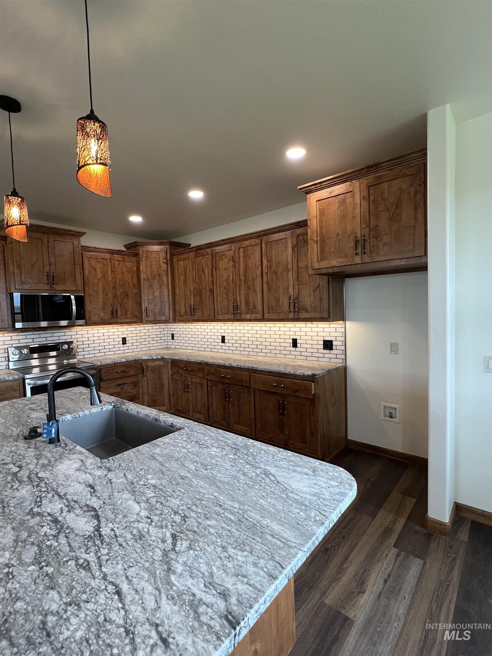 Kitchen featuring pendant lighting, brown cabinetry, dark wood-style flooring, backsplash, and appliances with stainless steel finishes