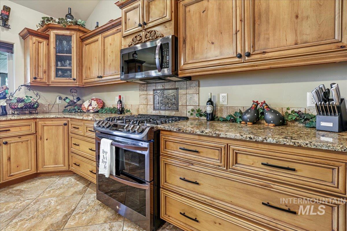 Kitchen with stainless steel appliances, tasteful backsplash, glass insert cabinets, light stone countertops, and brown cabinets