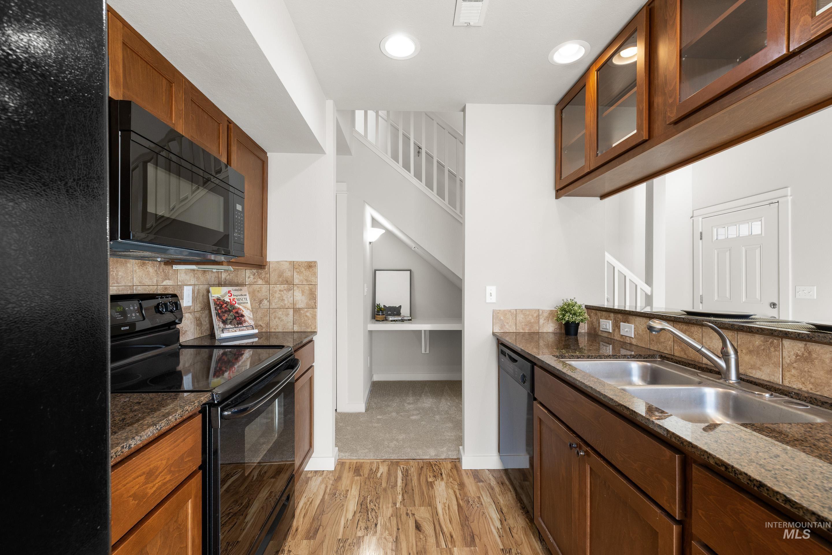 Kitchen with black appliances, light wood finished floors, glass fronted cabinets, dark stone counters, and backsplash