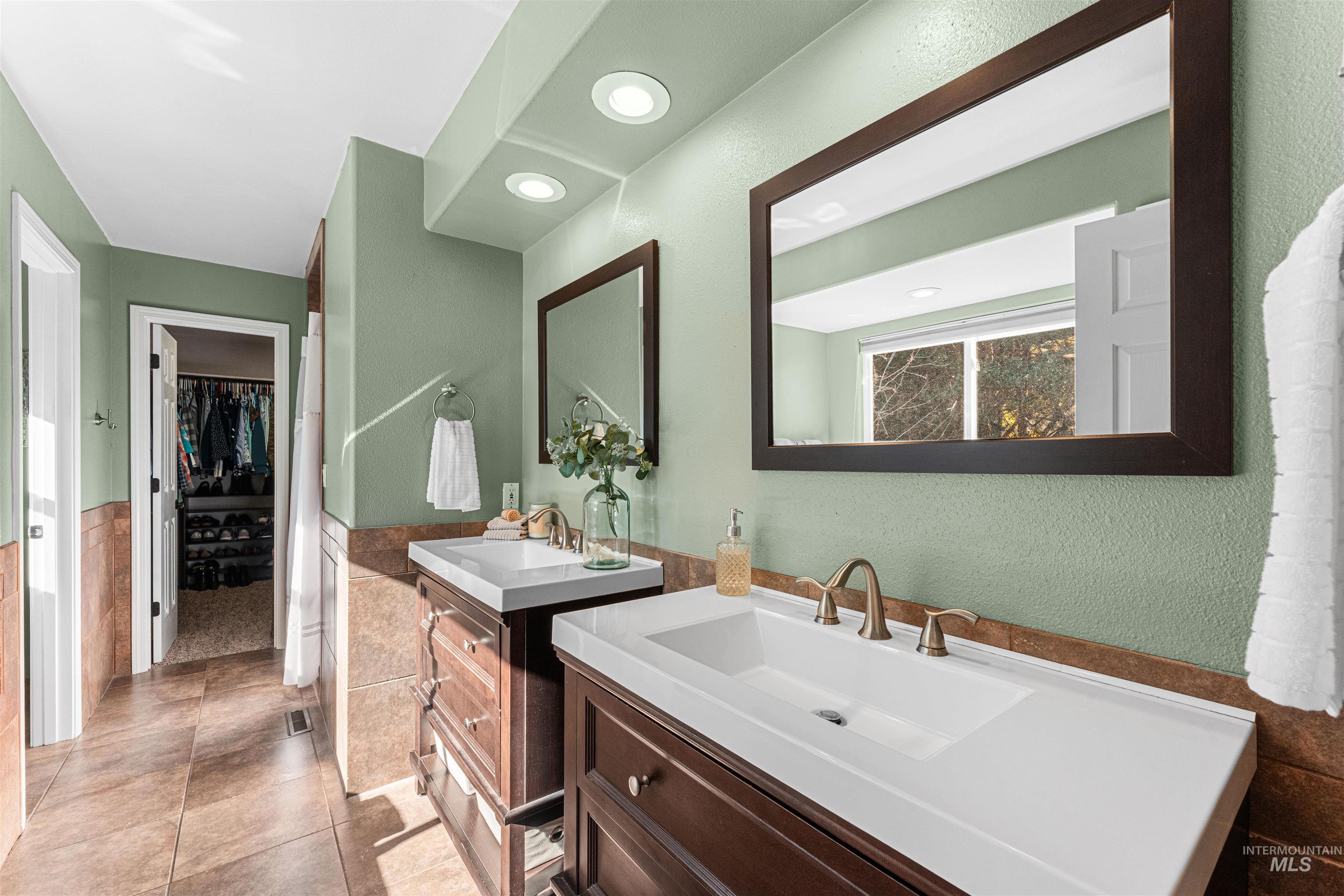 Full bath with two vanities, tile walls, light tile patterned flooring, a walk in closet, and wainscoting