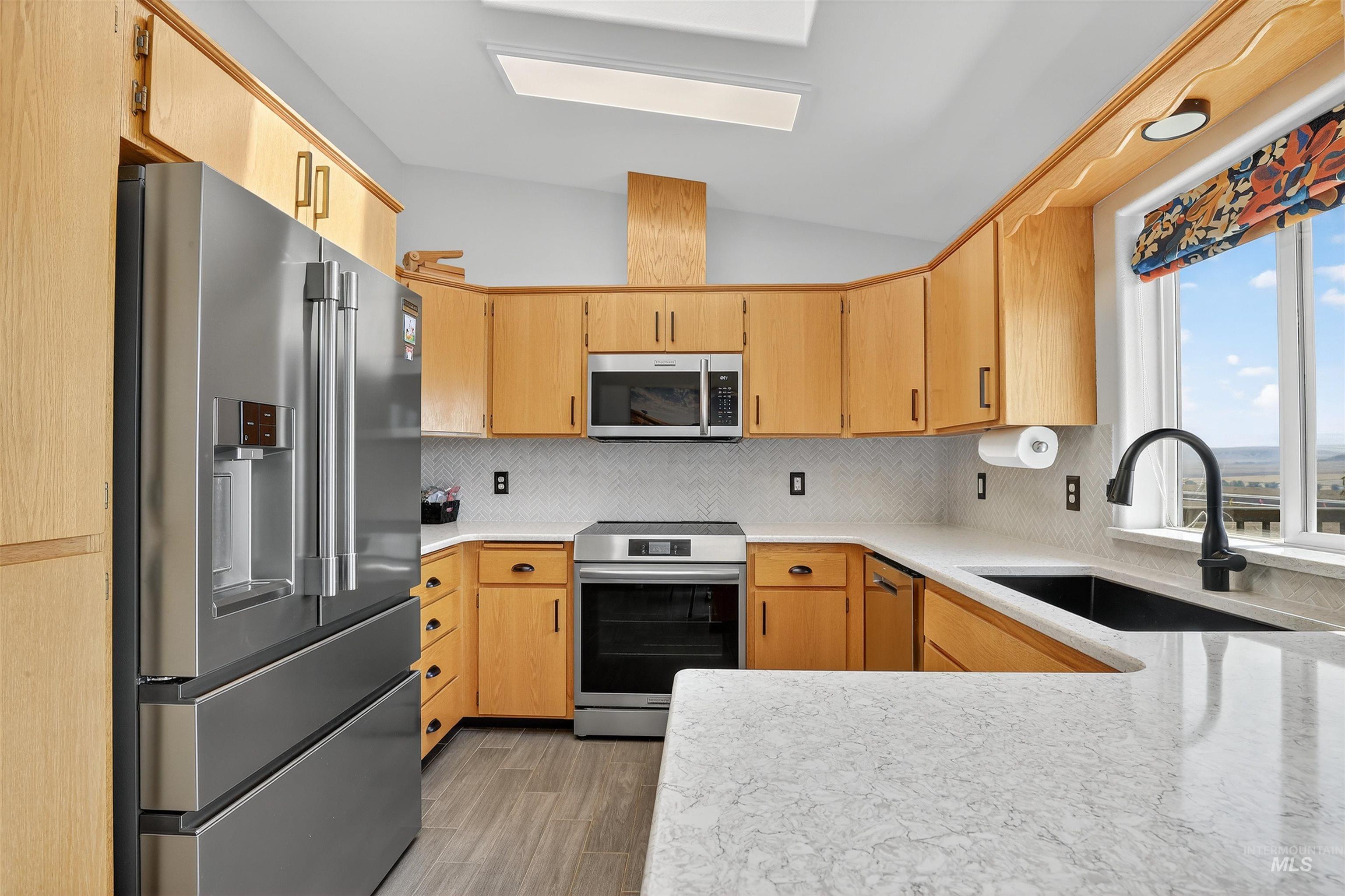 Kitchen featuring appliances with stainless steel finishes, decorative backsplash, light stone counters, light wood-style flooring, and light brown cabinetry